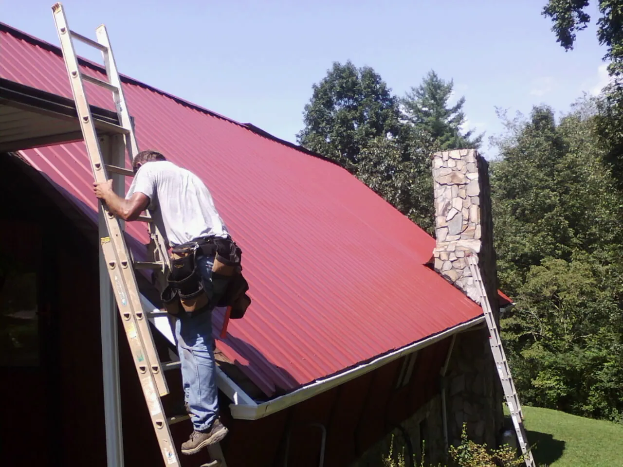 Roofer on a ladder beside a red metal roof and stone chimney — Soffit & Fascia Repair by Roofing Brothers in Union